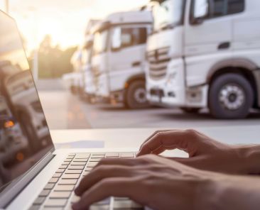 An image capturing a professional moment: close-up view of hands typing diligently on a laptop keyboard with the blur of transportation trucks in the sunlight creating a backdrop of industrious ambiance.